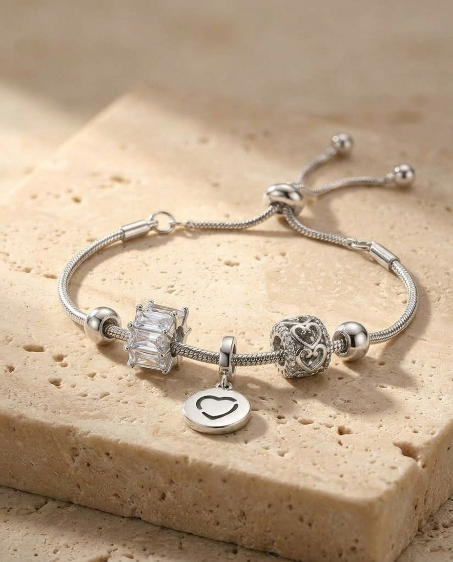 ALTUNA Love Charm Bracelet - silver snake chain with baguette, scrolling heart and open-heart disc charms, macro detail on travertine