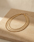 Camille Necklace editorial — layered gold chains on warm travertine