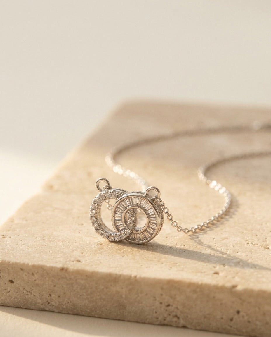 Claire Necklace by ALTUNA, macro low-angle detail of the interlocking pavé and baguette double-circle pendant on warm beige travertine
