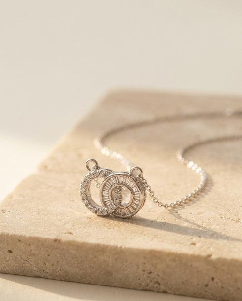 Claire Necklace by ALTUNA, macro low-angle detail of the interlocking pavé and baguette double-circle pendant on warm beige travertine