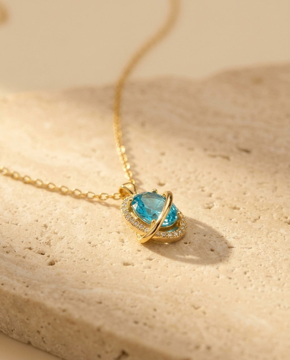 Nova Necklace detail — gold Saturn orbit pendant with faceted aquamarine stone and pave ring, low-angle macro on warm travertine