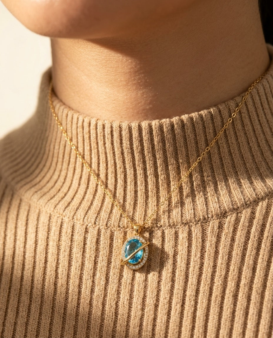 Nova Necklace worn — gold Saturn orbit pendant with aquamarine stone resting on a warm sand ribbed high-neck knit
