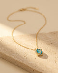 Nova Necklace editorial — gold orbit pendant with aquamarine stone on warm travertine