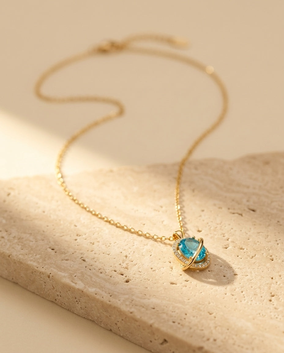 Nova Necklace editorial — gold orbit pendant with aquamarine stone on warm travertine