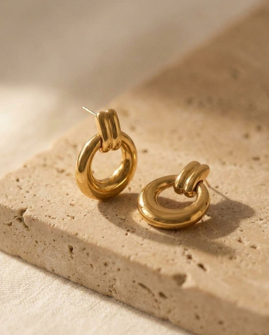 Oria Hoops by ALTUNA — angled studio view of the polished gold-tone drop hoops on warm travertine