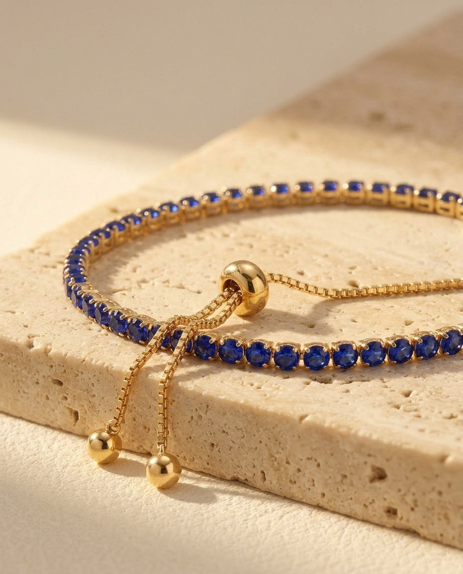 Seraphine Bracelet in Blue - macro three-quarter detail of the yellow gold tennis bracelet, royal blue stones and bolo slider on warm travertine