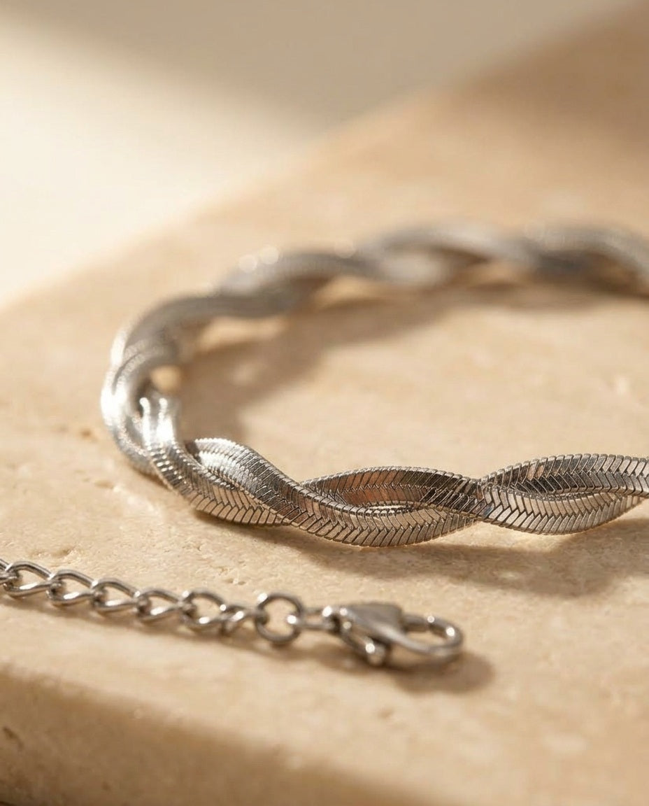Twisté Bracelet macro detail — two flat herringbone ribbons braided into a rope twist in polished silver, on warm beige travertine