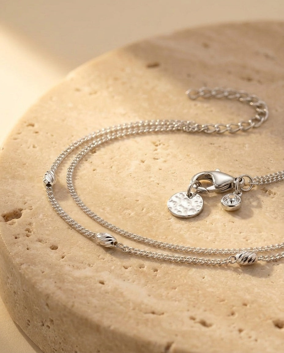 Véra Bracelet - macro three-quarter detail of the double-strand silver bracelet with twisted bead accents, hammered disc charm and bezel-set crystal on warm travertine