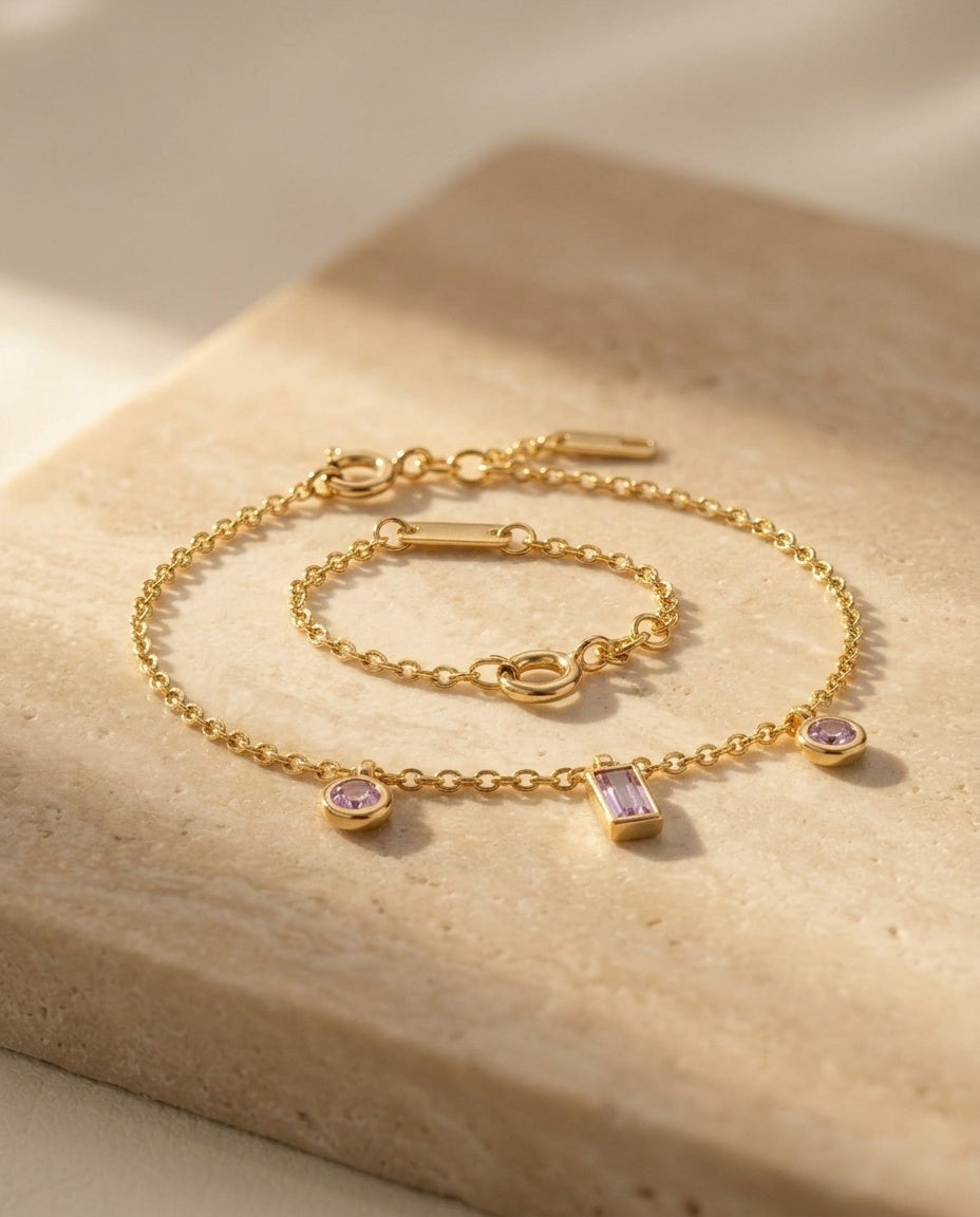 Violet Kiss Bracelet — macro three-quarter detail of the gold chain and pale violet amethyst charms on travertine